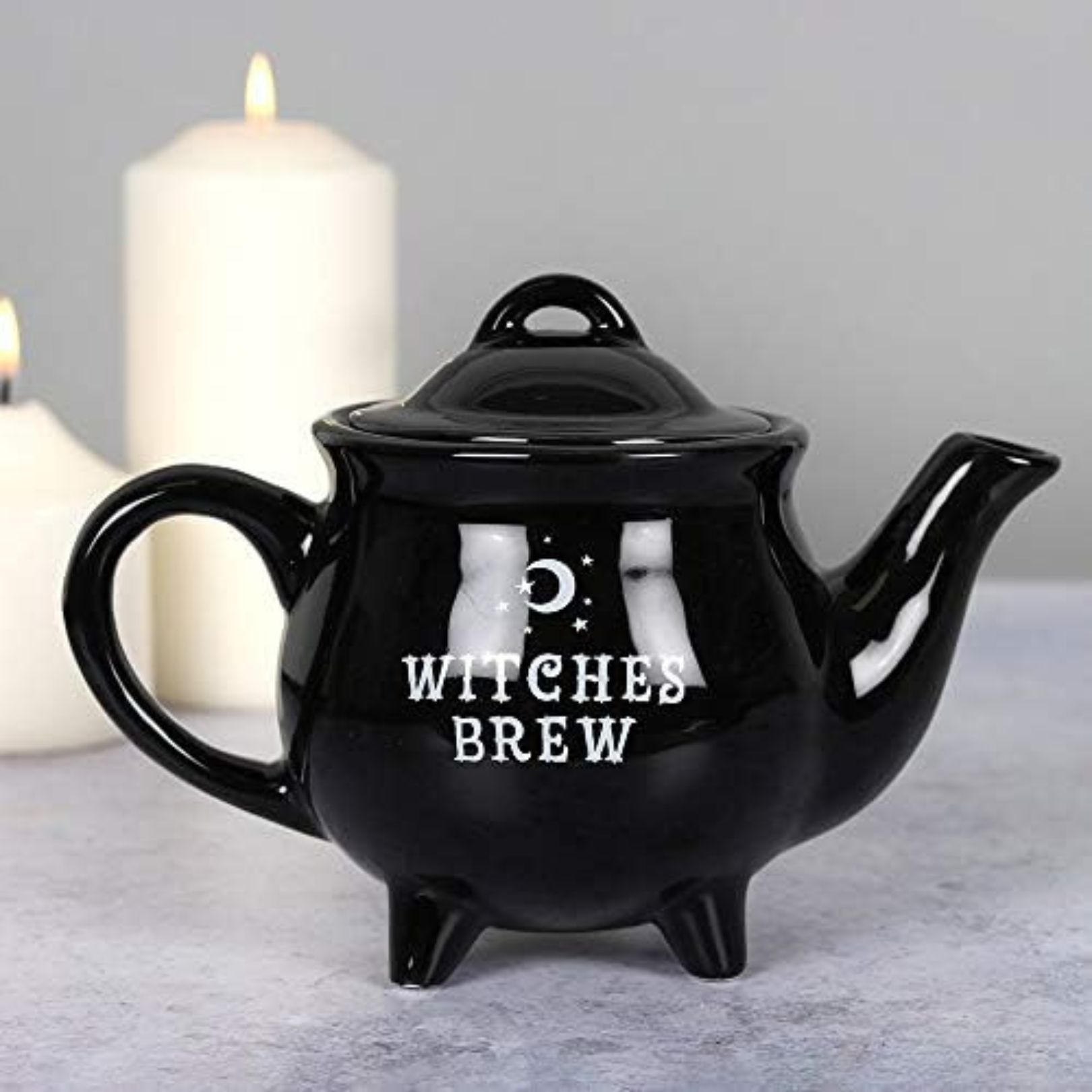Classic black teapot with witches brew printed in white on the side, in front of a white pillar candle giving a witchy decor look