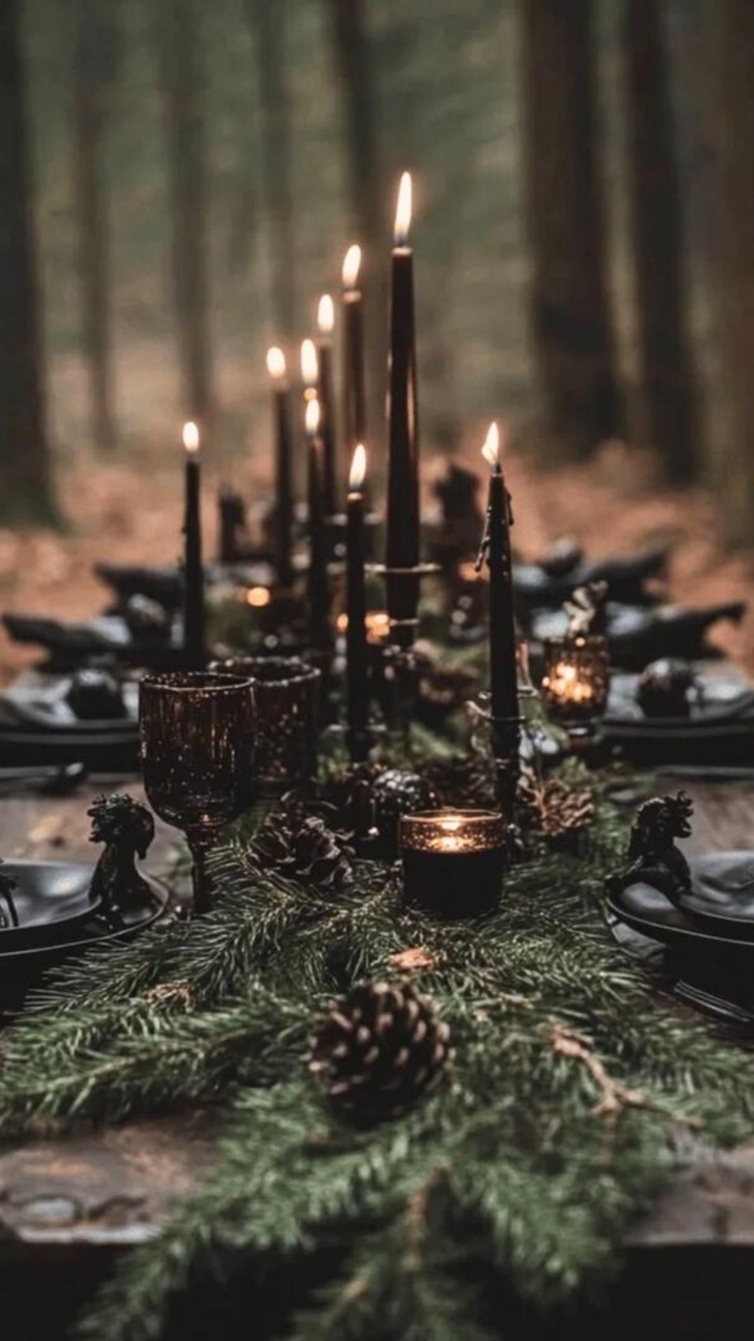 Moody forest witch tablescape with black candles and smokey glass goblets, set outside in a pine forest